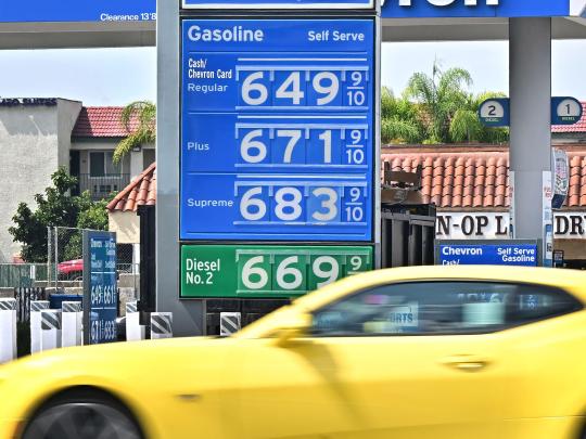 A yellow car driving in front of a gas station with gas prices starting at $6.49 per gallon.