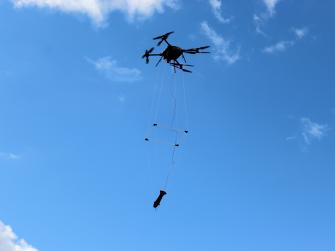 A drone flies through a blue sky with clouds carrying a magnetometer suspended below it.